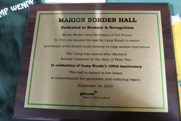 Gold plaque with wood trim that reads "Marion Borden Hall Dedicated in Memory & Recognition. Marion Boarden loved the mission of Girl Scouts. In 1923, she donated the land for Camp Wendy to ensure generations of Girl Scouts council continue to enjoy outdoor experiences. The Camp was names after Marion's favorite character in the story of Peter Pan. In celebration of Camp Wendy's 100th Anniversary this hall is named in her honor to commemorate her generosity and enduring legacy. 